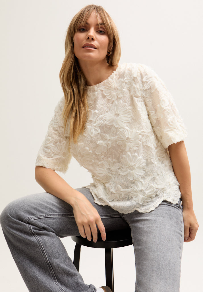Woman wearing a white textured top and gray jeans sitting on a black stool against a light background