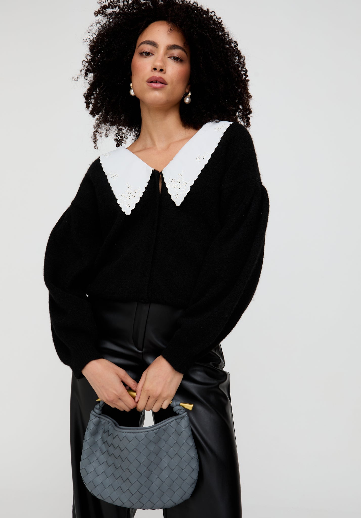 Woman wearing a black outfit with a white collar holding a gray handbag against a white background