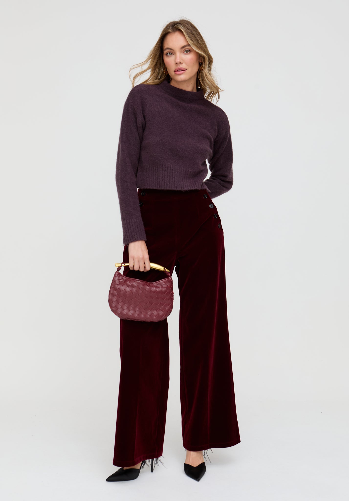 Woman wearing a dark purple sweater and pants, holding a matching handbag on a white background
