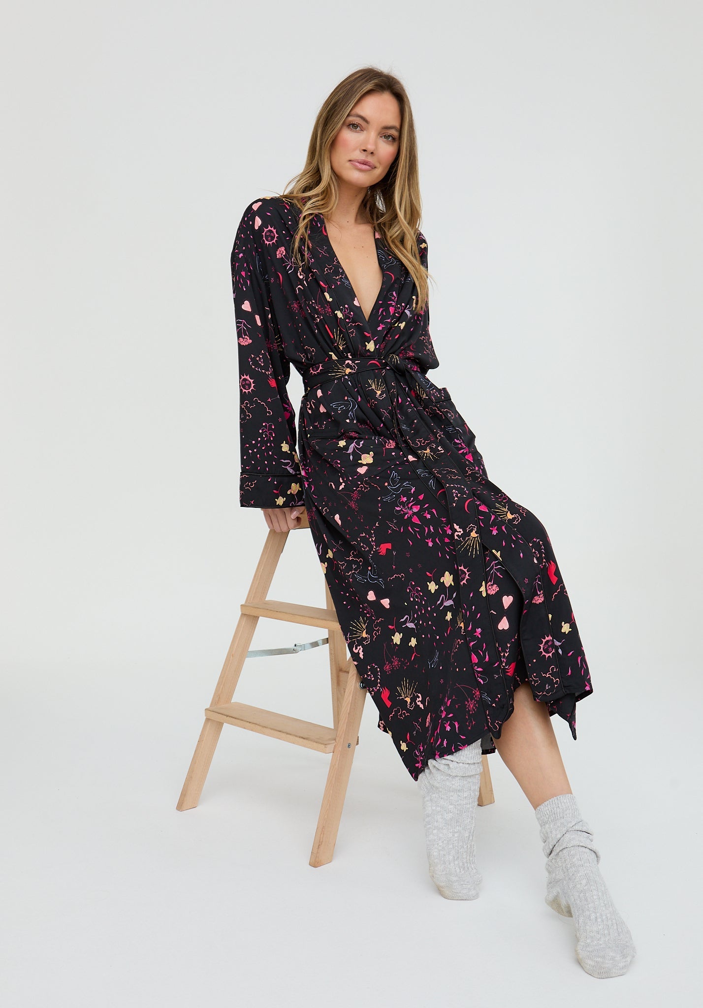 Woman wearing a black floral dress sitting on a wooden stool against a white background