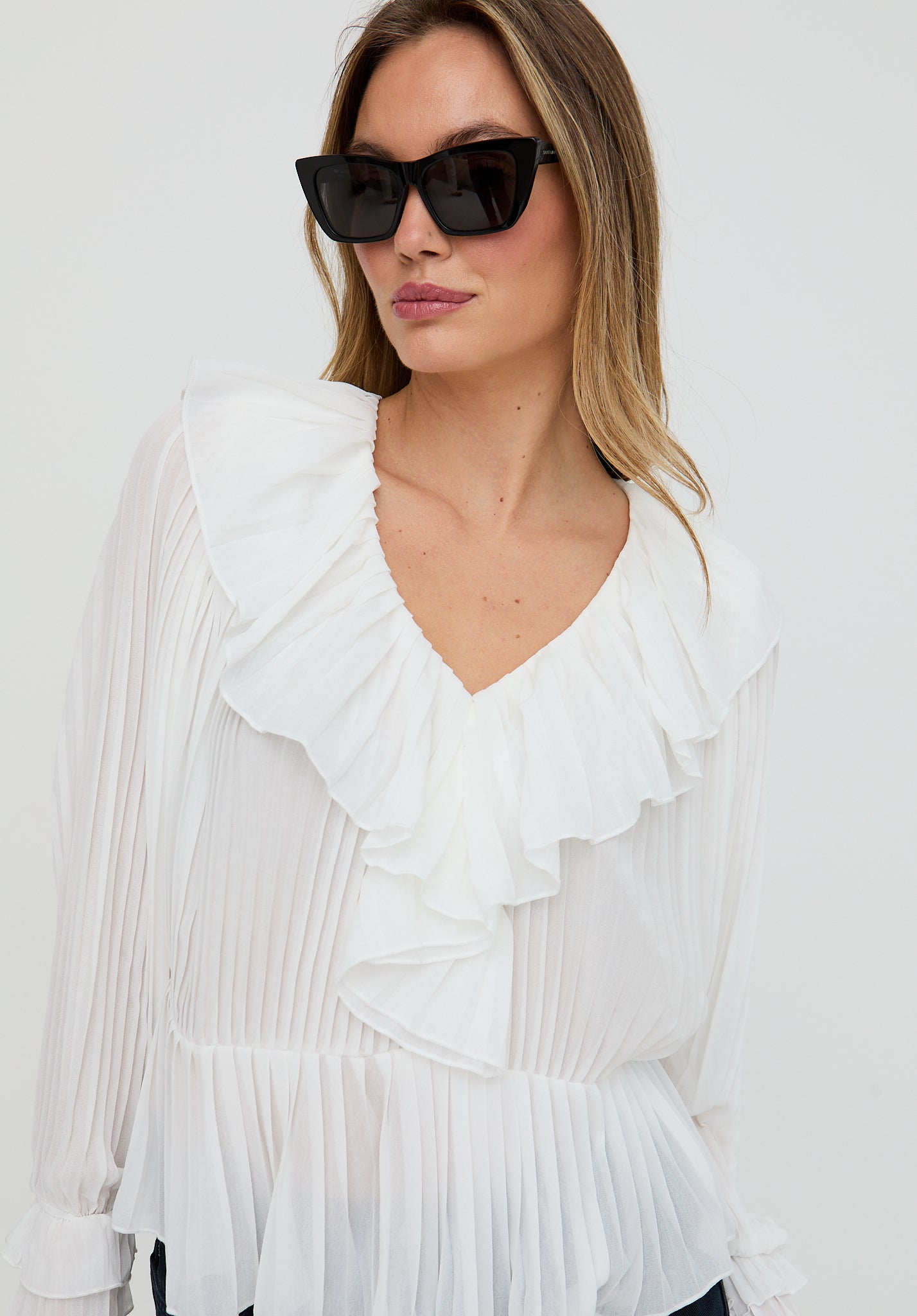 Woman wearing a white pleated blouse with ruffles and black sunglasses against a white background