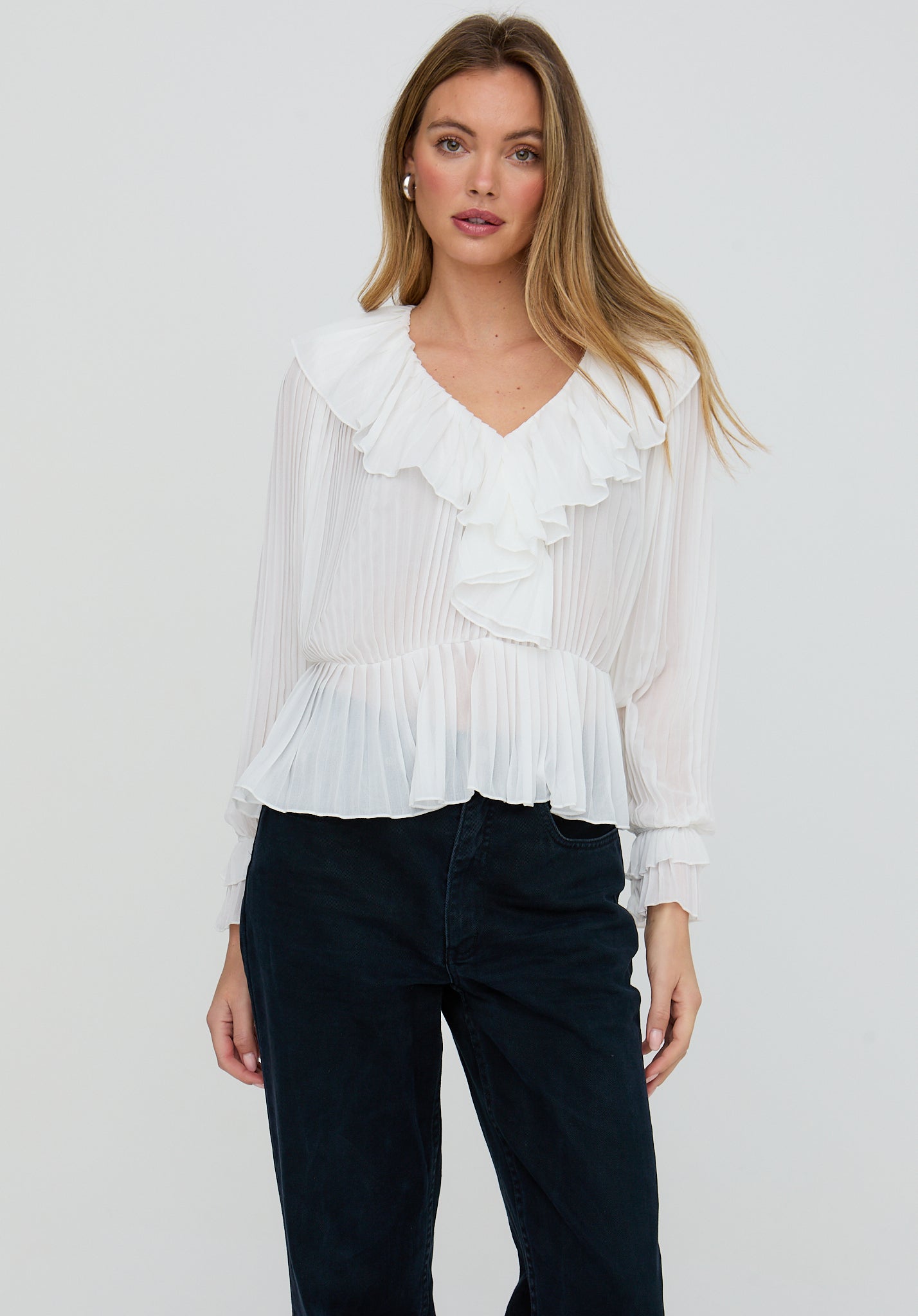 Woman wearing a white blouse with ruffles and dark pants on a plain background