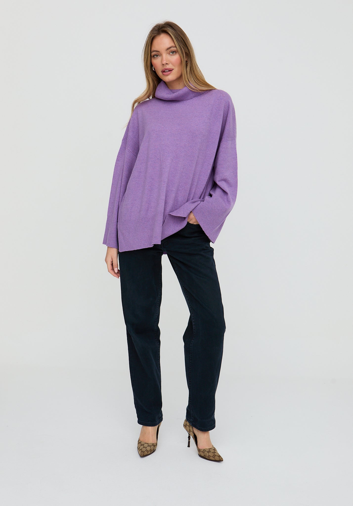 Woman wearing a purple cowl-neck sweater and black pants on a white background
