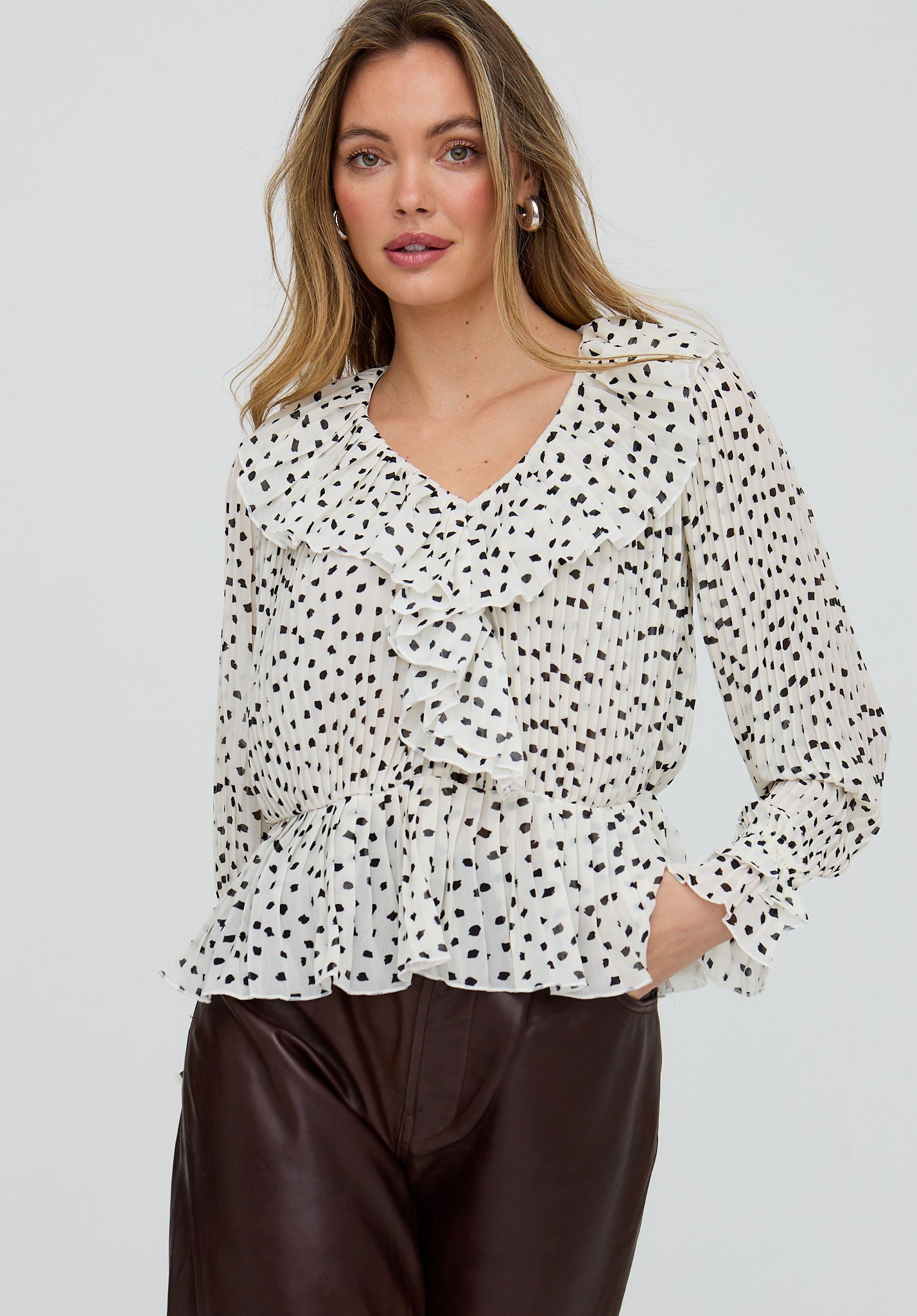 Woman wearing a white blouse with black polka dots and brown pants on a light gray background