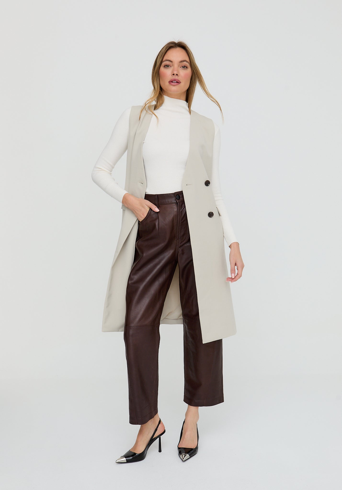 Woman wearing a long beige coat over a white top and brown pants on a white background