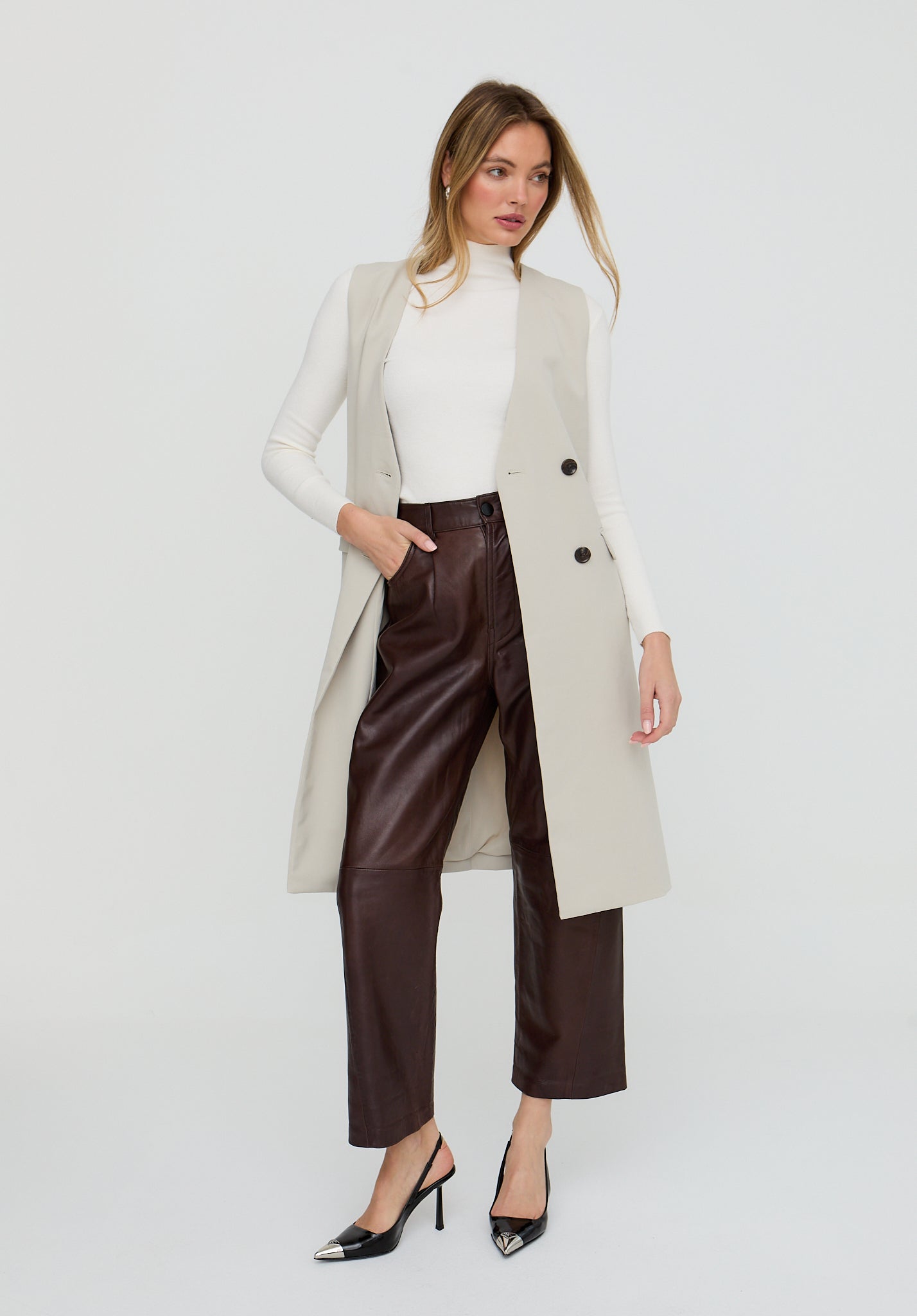 Woman wearing a beige long coat, white top, and brown pants on a light gray background