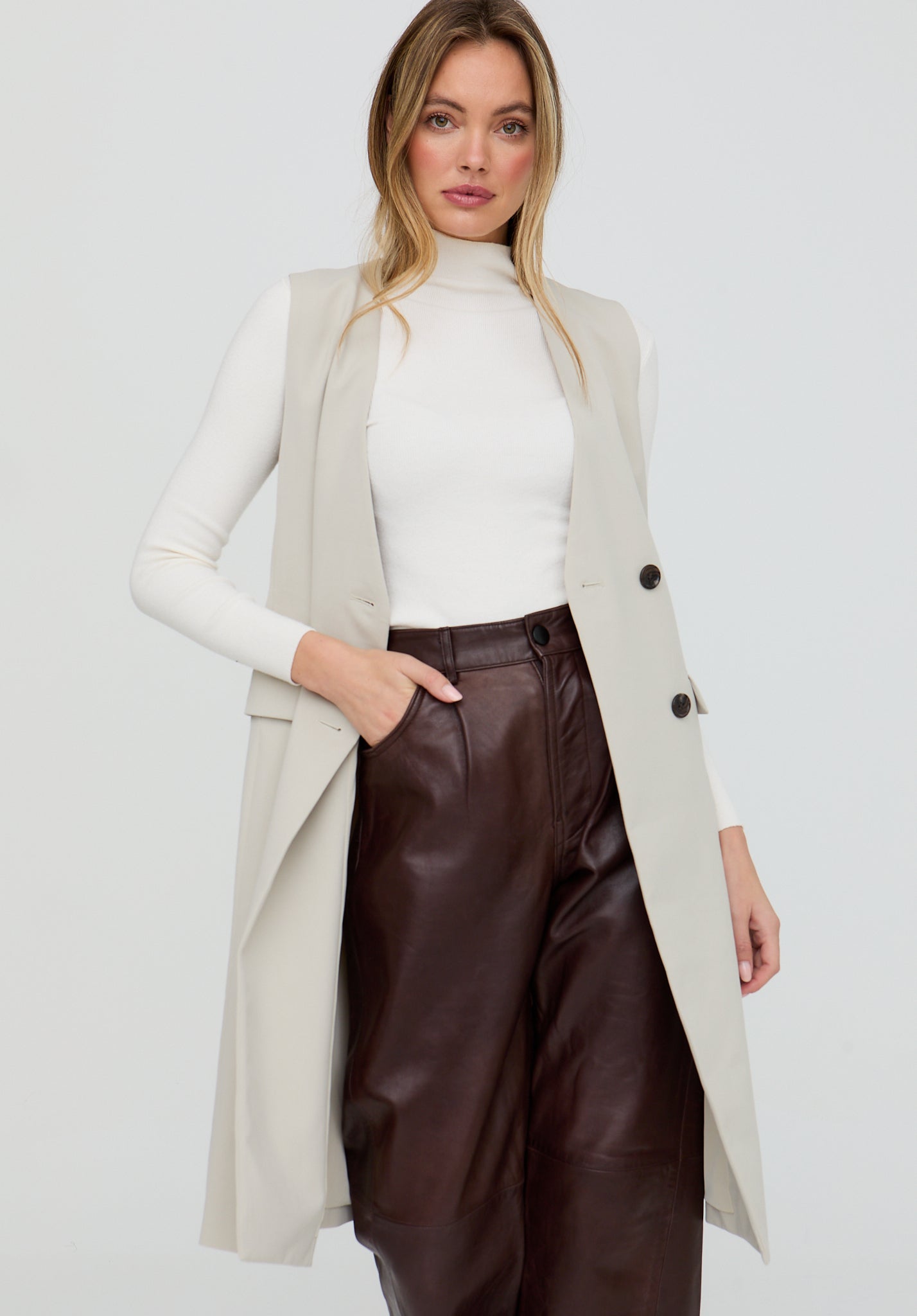 Woman wearing a long beige coat over a white turtleneck and brown leather pants on a light gray background