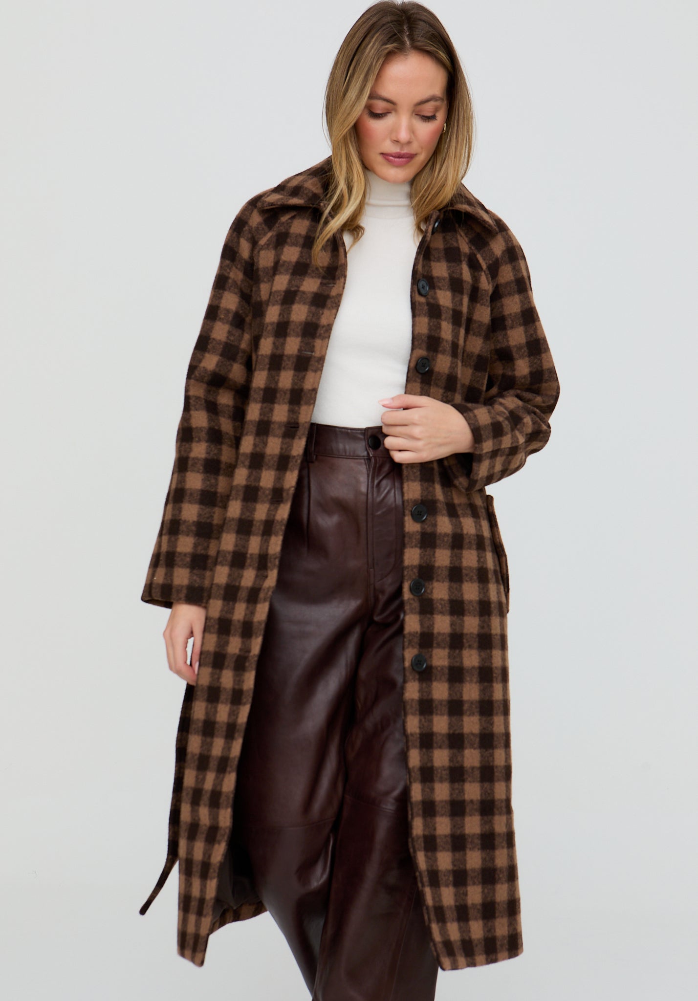Woman wearing a brown plaid coat over a white top and brown leather pants on a light gray background