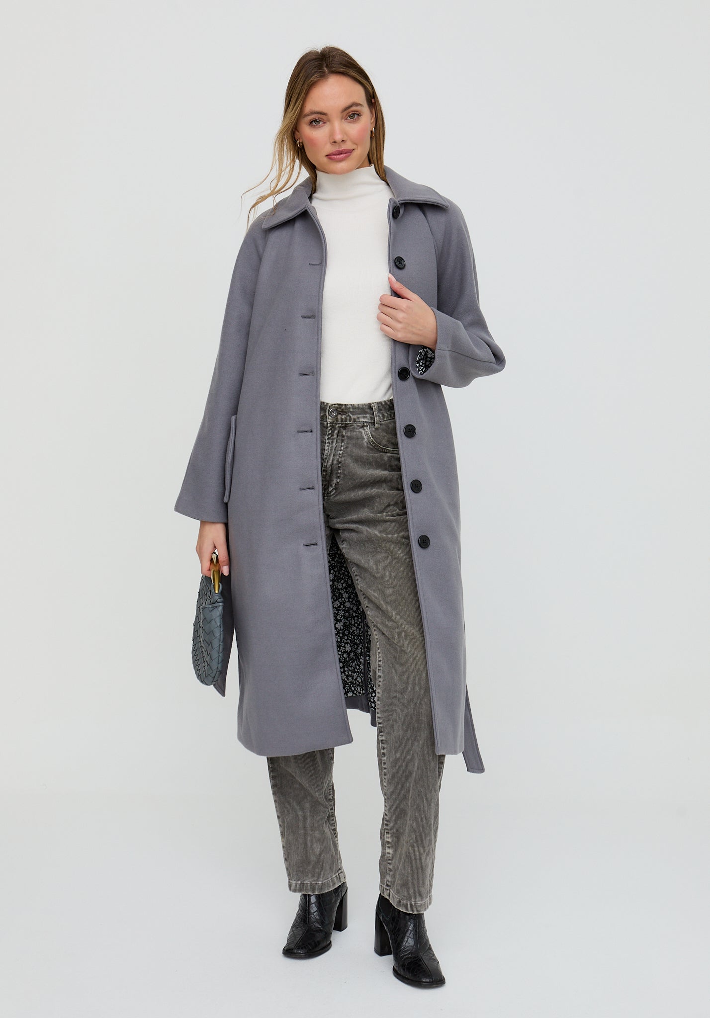 Woman wearing a gray coat, white top, and green pants on a light gray background