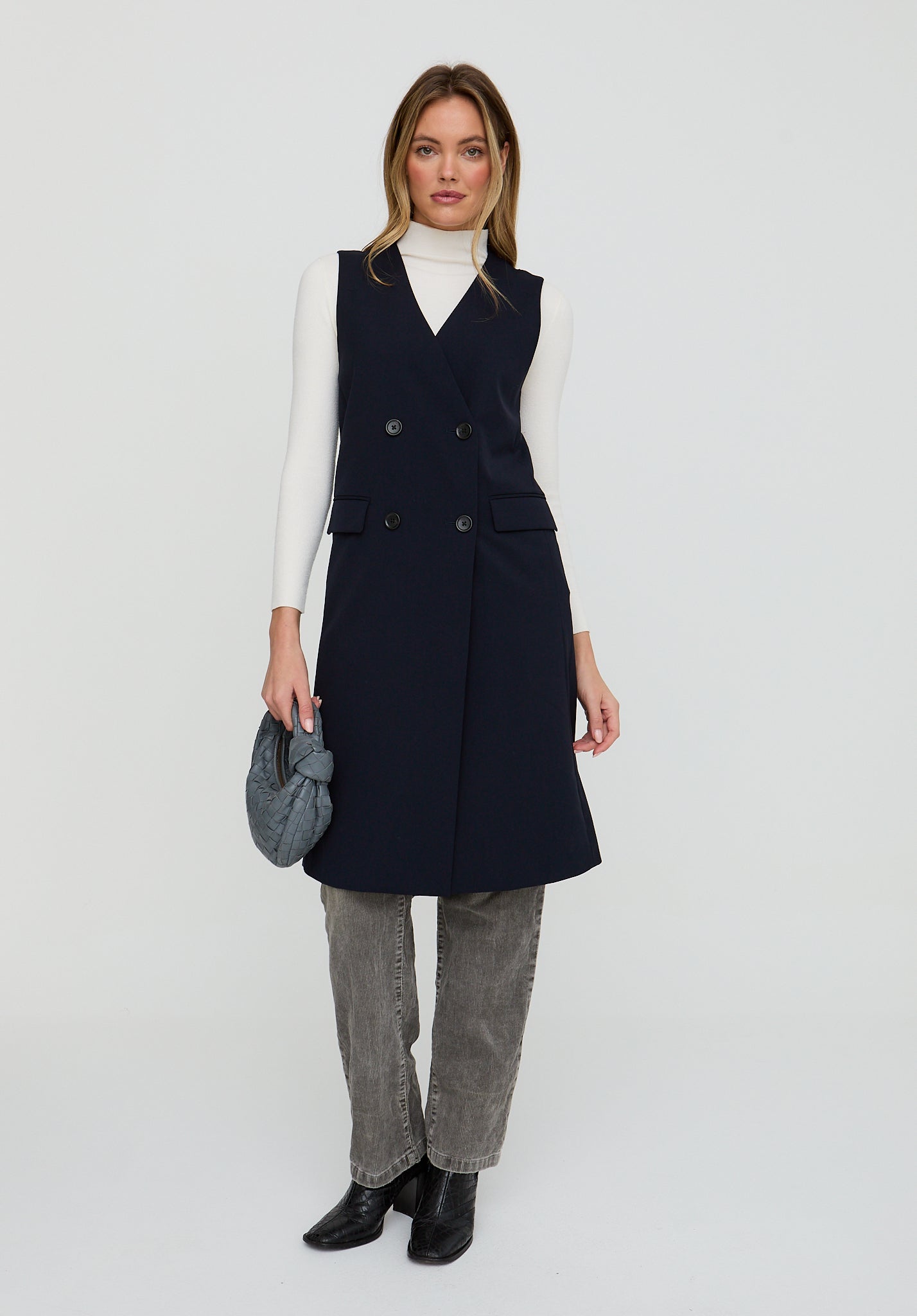 Woman wearing a navy blue dress over a white turtleneck with gray jeans and black shoes on a white background