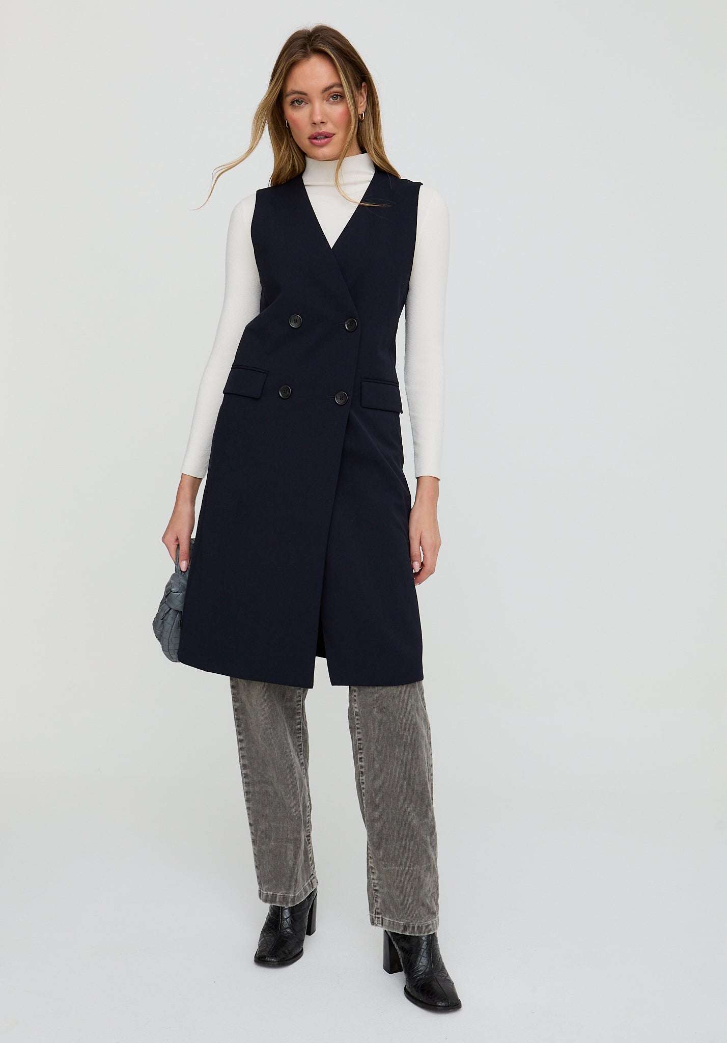 Woman wearing a black vest over a white turtleneck with gray pants on a light gray background