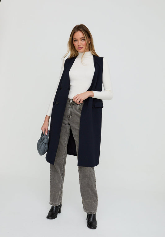 Woman wearing a long navy vest over a white turtleneck and gray jeans on a white background