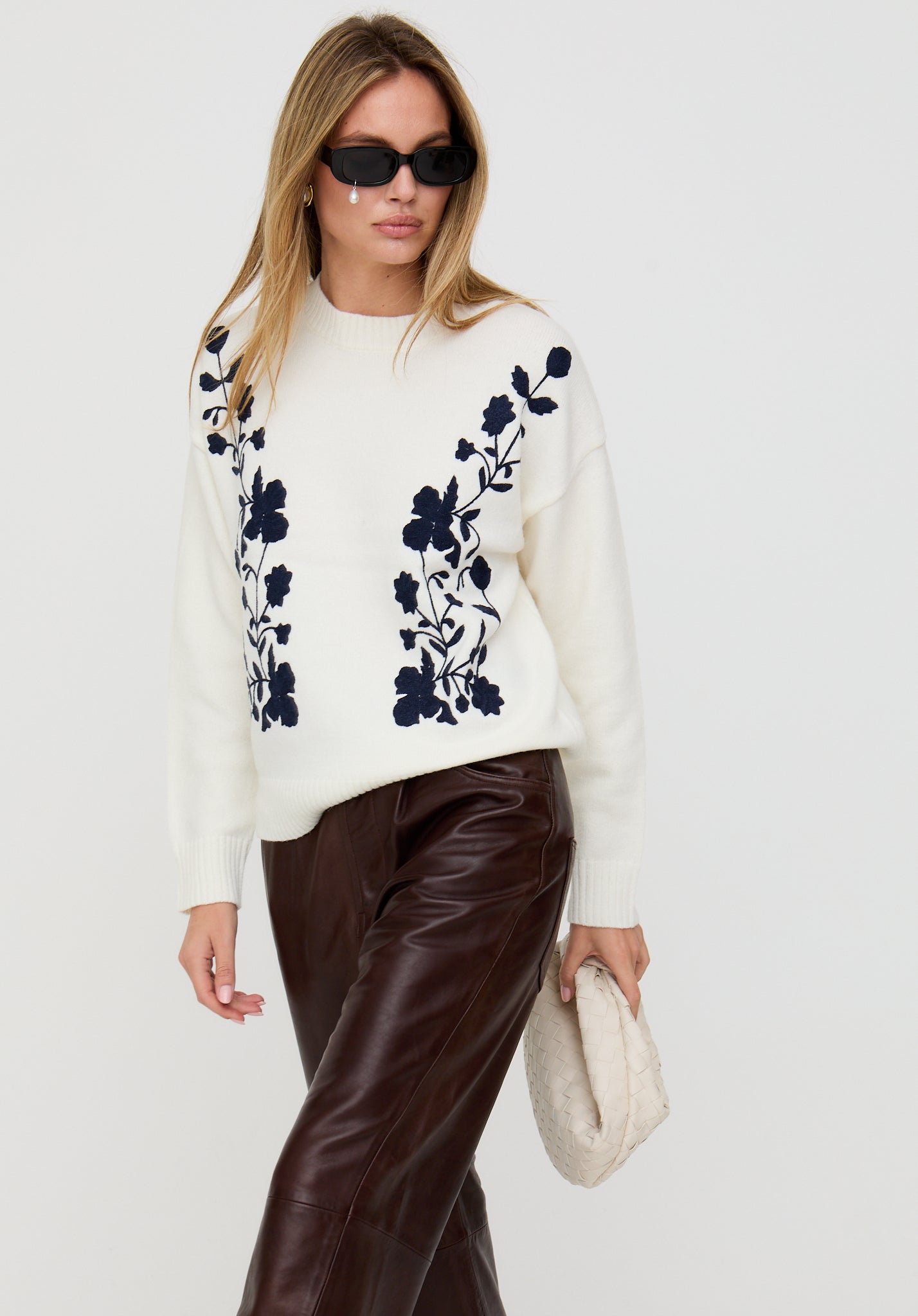 Woman wearing a white sweater with black floral patterns, brown pants, and sunglasses on a light gray background.