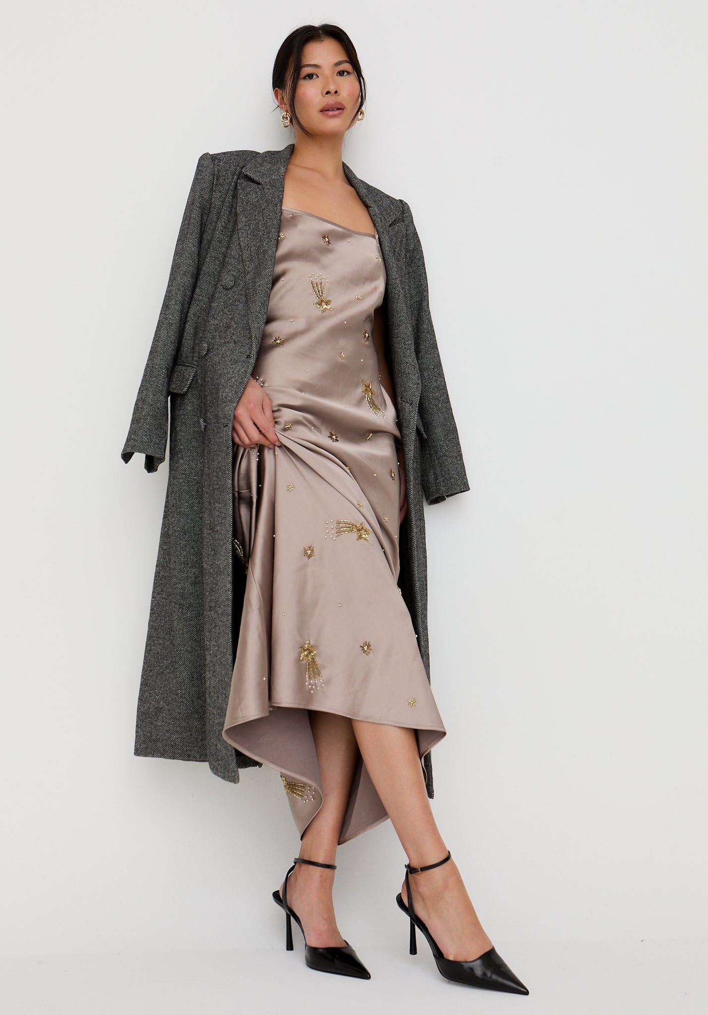 Woman wearing a long gray coat over a beige dress with floral embroidery on a white background