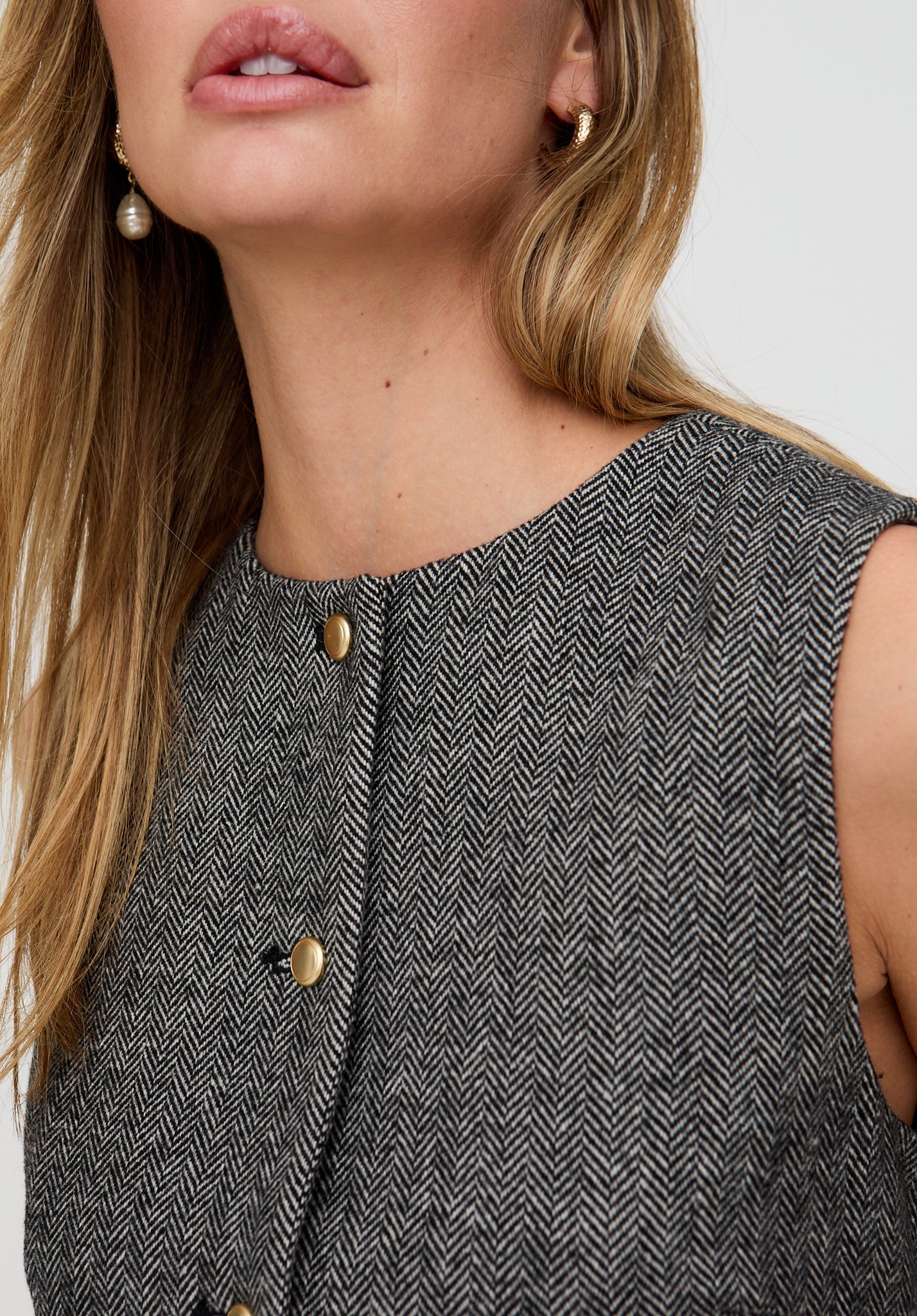 Woman wearing a gray herringbone patterned sleeveless top with gold buttons.