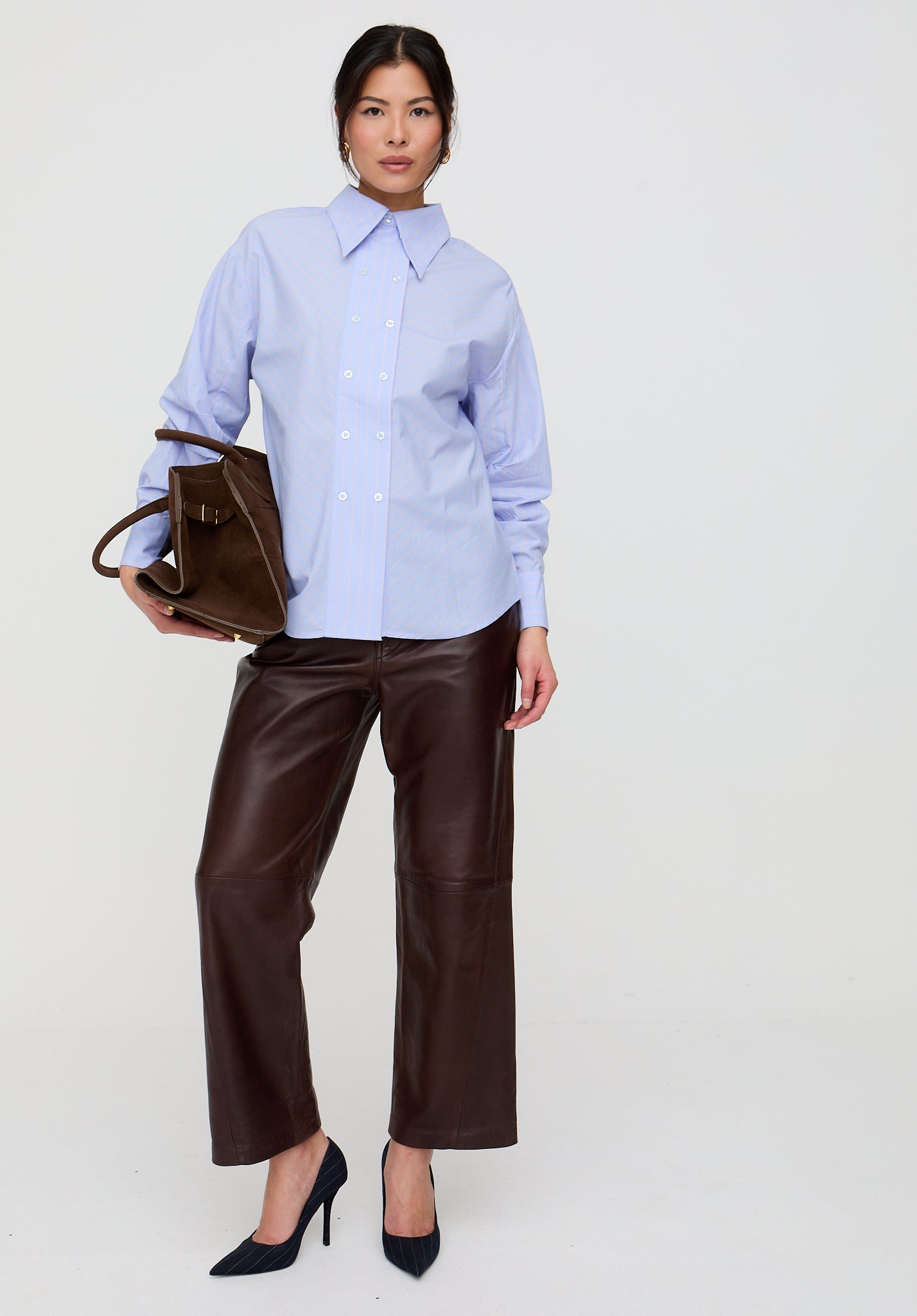 Woman wearing a light blue shirt and brown leather pants, holding a brown leather bag on a white background
