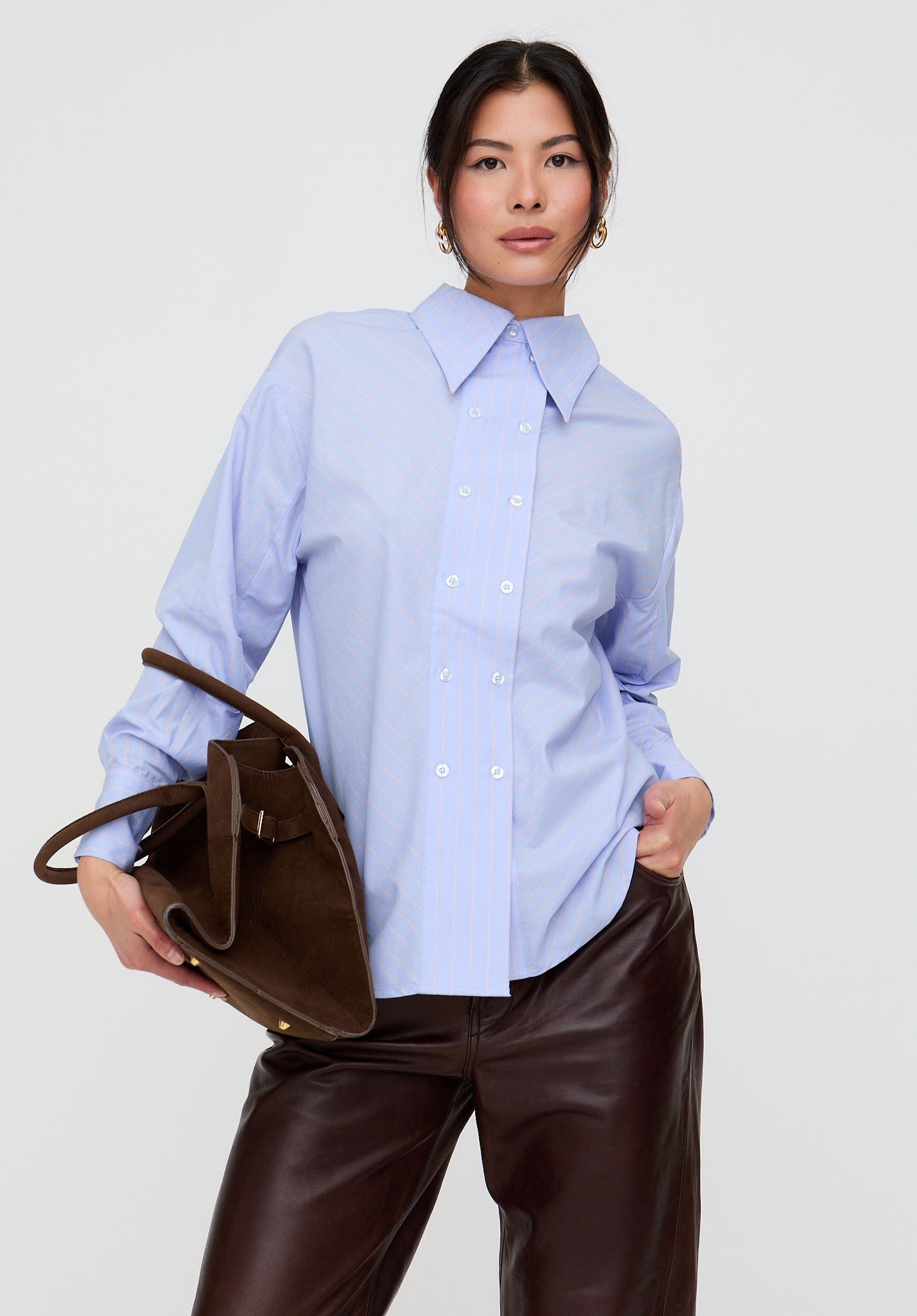 Woman wearing a light blue shirt and brown pants holding a brown handbag on a white background
