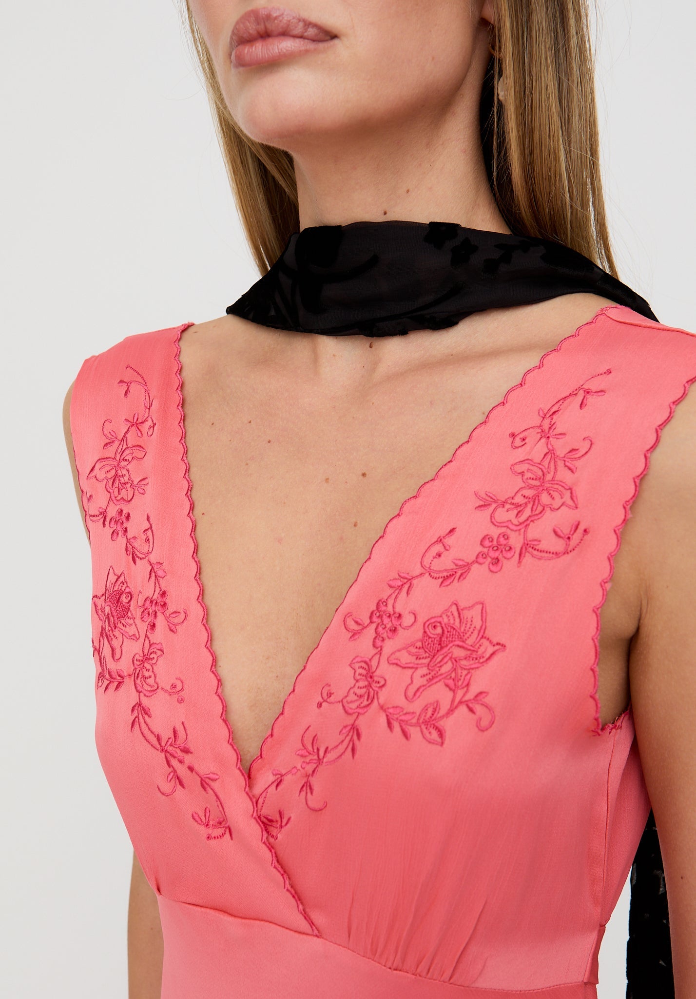 Close-up of a pink dress with floral embroidery and a black scarf around the neck on a white background.