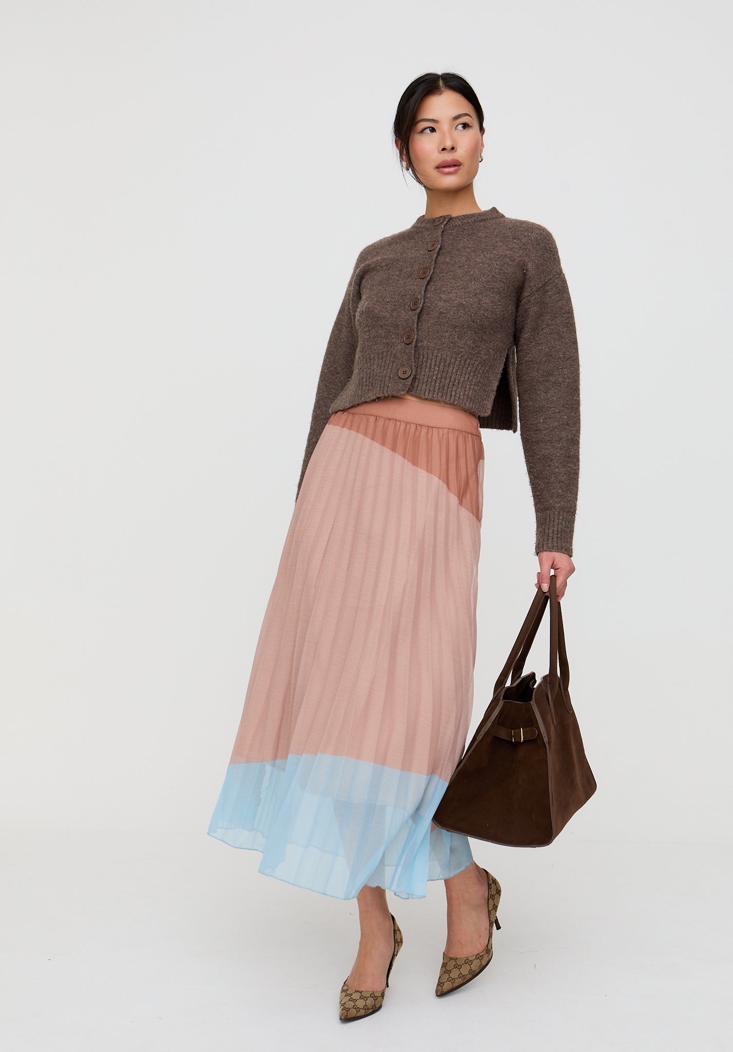 Woman wearing a brown cardigan and a pleated skirt with a gradient effect, holding a brown handbag on a white background.