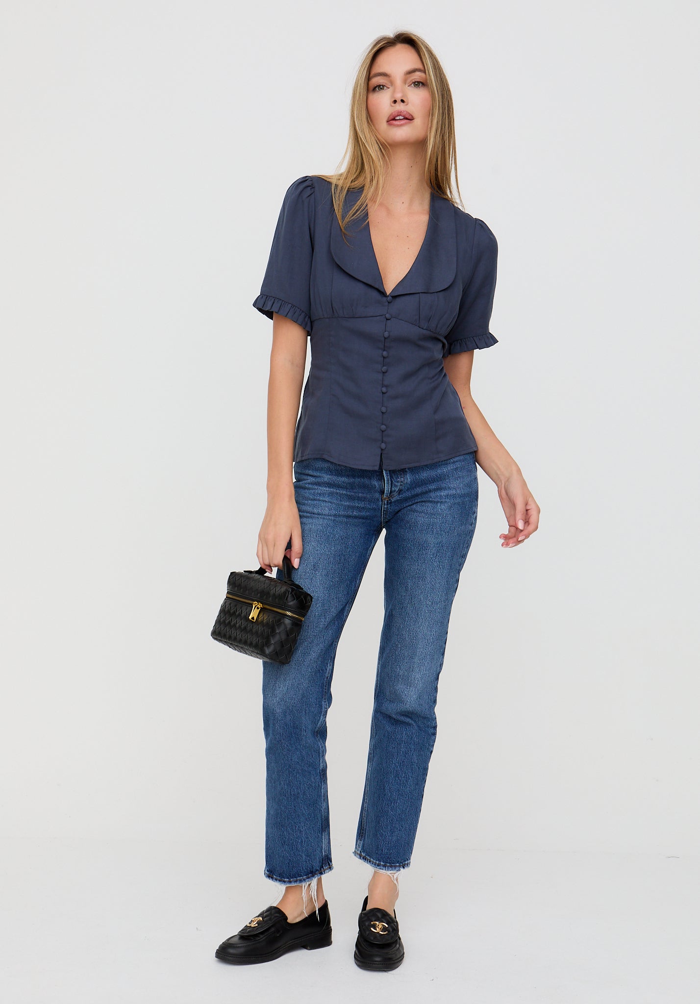 Woman wearing a navy blue top and jeans holding a black handbag on a white background